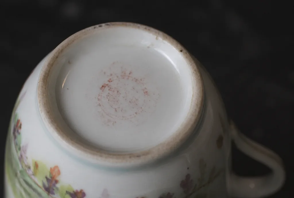 AI illustration: the underside of a small hand-painted porcelain teacup turned upside down, a faint red-brown printed mark on the white base, the lettering too faded to read